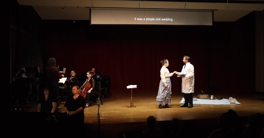On left, a chamber orchestra, conductor, and two women doing ASL interpretation. On right, YOUNG MARIE (Alishia Garcia) and PIERRE CURIE (Zachary Lenox)  sing together.